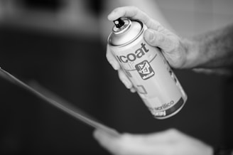 A hand is holding and using an aerosol spray can, applying it to an unseen surface. The can has visible branding and text.