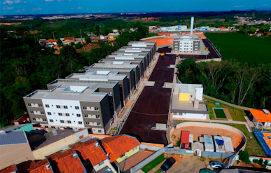 An aerial view of a residential area under development. Multiple multi-story apartment buildings dominate the scene, aligned in a row along a newly constructed road. Surrounding these structures are patches of green forested areas and open fields. A mix of construction sites and completed buildings is visible, along with some smaller houses with orange rooftops. The landscape extends into the distance with clear blue skies above.