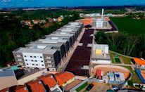 An aerial view of a residential area under development. Multiple multi-story apartment buildings dominate the scene, aligned in a row along a newly constructed road. Surrounding these structures are patches of green forested areas and open fields. A mix of construction sites and completed buildings is visible, along with some smaller houses with orange rooftops. The landscape extends into the distance with clear blue skies above.