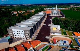 An aerial view of a residential area under development. Multiple multi-story apartment buildings dominate the scene, aligned in a row along a newly constructed road. Surrounding these structures are patches of green forested areas and open fields. A mix of construction sites and completed buildings is visible, along with some smaller houses with orange rooftops. The landscape extends into the distance with clear blue skies above.