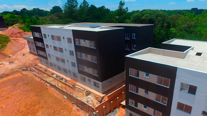 Aerial view of a partially completed multi-story building complex with modern architecture, featuring a mix of black and white facades. The construction site is surrounded by a natural environment with dense greenery and trees in the background. There are sections of exposed earth and construction materials visible along the perimeter.