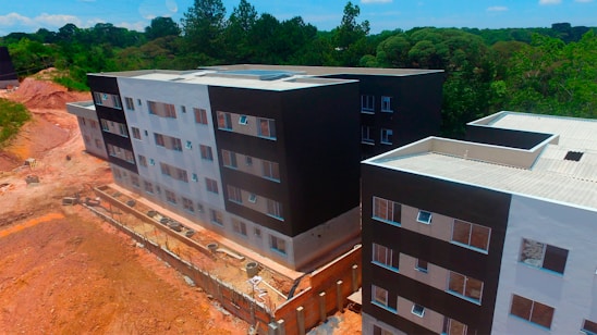 Aerial view of a partially completed multi-story building complex with modern architecture, featuring a mix of black and white facades. The construction site is surrounded by a natural environment with dense greenery and trees in the background. There are sections of exposed earth and construction materials visible along the perimeter.