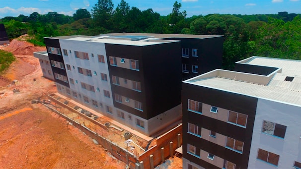 Aerial view of a partially completed multi-story building complex with modern architecture, featuring a mix of black and white facades. The construction site is surrounded by a natural environment with dense greenery and trees in the background. There are sections of exposed earth and construction materials visible along the perimeter.
