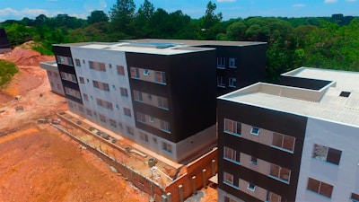 Aerial view of a partially completed multi-story building complex with modern architecture, featuring a mix of black and white facades. The construction site is surrounded by a natural environment with dense greenery and trees in the background. There are sections of exposed earth and construction materials visible along the perimeter.