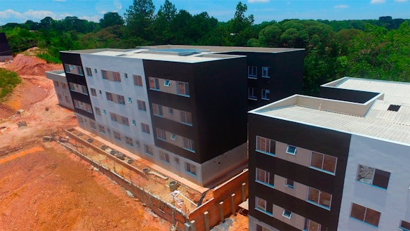 Aerial view of a partially completed multi-story building complex with modern architecture, featuring a mix of black and white facades. The construction site is surrounded by a natural environment with dense greenery and trees in the background. There are sections of exposed earth and construction materials visible along the perimeter.