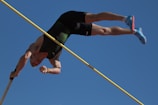 An athlete is in mid-air performing a pole vault against a clear blue sky. The athlete is upside down with one hand gripping the pole and their legs extended upwards. They are wearing a dark athletic outfit and bright blue shoes.