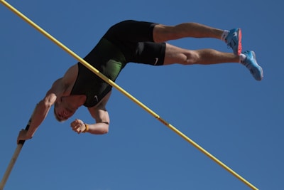 An athlete is in mid-air performing a pole vault against a clear blue sky. The athlete is upside down with one hand gripping the pole and their legs extended upwards. They are wearing a dark athletic outfit and bright blue shoes.