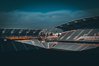 A large, empty sports stadium featuring rows of seats in the stands. The architecture includes a modern roof structure and is partially shaded. In the background, a skyline with buildings and palm trees is visible under a cloudy sky. There is a glowing sign with text in Spanish suggesting a connection to the local area.