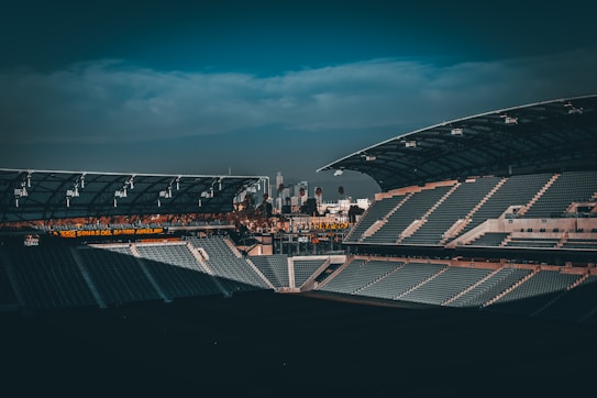 A large, empty sports stadium featuring rows of seats in the stands. The architecture includes a modern roof structure and is partially shaded. In the background, a skyline with buildings and palm trees is visible under a cloudy sky. There is a glowing sign with text in Spanish suggesting a connection to the local area.