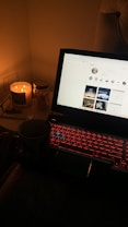 A dimly lit room featuring a laptop with a red backlit keyboard displaying a social media page. A lit candle and a jar labeled 'NICHES' are placed beside the laptop, with a hand holding a mug visible in the foreground.