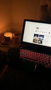 A dimly lit room featuring a laptop with a red backlit keyboard displaying a social media page. A lit candle and a jar labeled 'NICHES' are placed beside the laptop, with a hand holding a mug visible in the foreground.