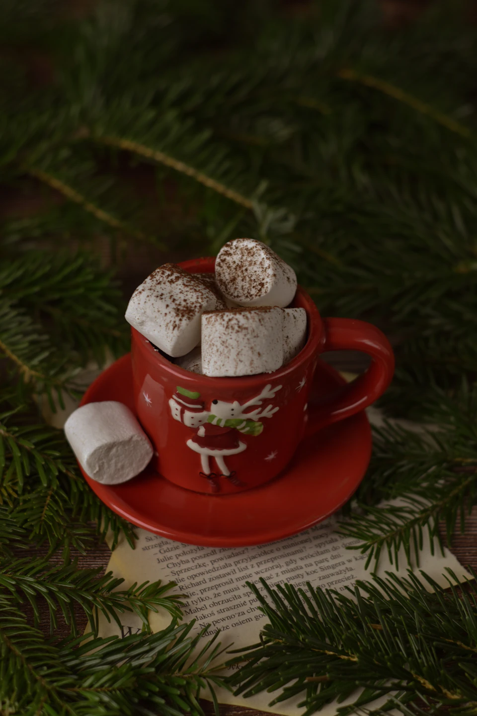 The image presents a festive scene set against a backdrop of Christmas trees and greenery. Dominating the center is a red mug, adorned with a white design featuring a reindeer holding a snowflake. The mug is filled to the brim with small marshmallows, their white color contrasting beautifully with the red of the mug. To the right of the mug, there's a stack of papers, possibly containing holiday greetings or other festive messages.