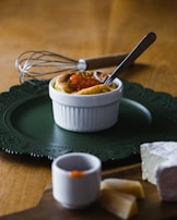 A small white ramekin containing a baked dish topped with a small amount of orange sauce is placed on a dark green decorative plate. A metal whisk with a wooden handle is in the background. In the foreground on a wooden board, there is a piece of brie cheese with two slices next to it and a small cup with some sauce on the rim. A silver spoon rests inside the ramekin.