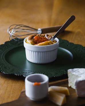 A small white ramekin containing a baked dish topped with a small amount of orange sauce is placed on a dark green decorative plate. A metal whisk with a wooden handle is in the background. In the foreground on a wooden board, there is a piece of brie cheese with two slices next to it and a small cup with some sauce on the rim. A silver spoon rests inside the ramekin.