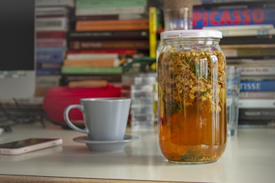 A glass jar filled with a light brown liquid containing herbs or flowers is placed on a desk. Around it are a ceramic cup and saucer, a smartphone, and stacks of books in the background, giving a cozy and cluttered work or study space vibe.