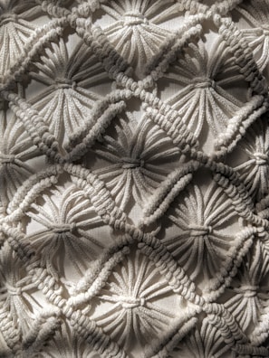 Intricate macrame pattern showcasing a series of interconnected diamond shapes, each formed by tightly knotted and looped threads. The texture is richly detailed, with depth created by the overlapping threads casting subtle shadows.