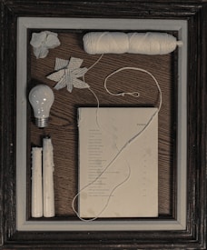 The image features a framed collection of objects neatly arranged on a wooden surface. Items include a light bulb, two white candles, a ball of white string, a piece of paper with printed text, and a decorative paper star. A white flower appears in the corner. The overall aesthetic suggests a vintage or rustic theme.
