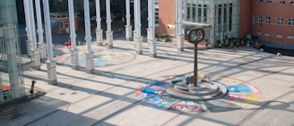 A spacious courtyard with a circular tiled artwork on the ground, featuring vibrant colors and various designs. In the center stands a tall, unique sculpture resembling a clock or compass. Multiple white columns cast shadows across the area, and a large building with glass windows and red brick walls surrounds the courtyard.