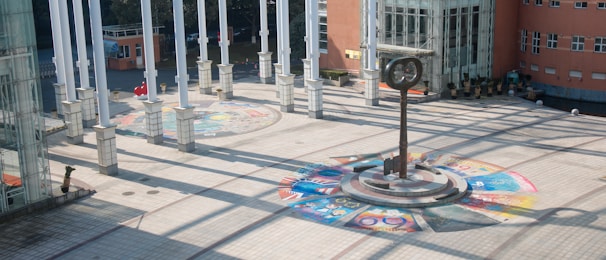 A spacious courtyard with a circular tiled artwork on the ground, featuring vibrant colors and various designs. In the center stands a tall, unique sculpture resembling a clock or compass. Multiple white columns cast shadows across the area, and a large building with glass windows and red brick walls surrounds the courtyard.
