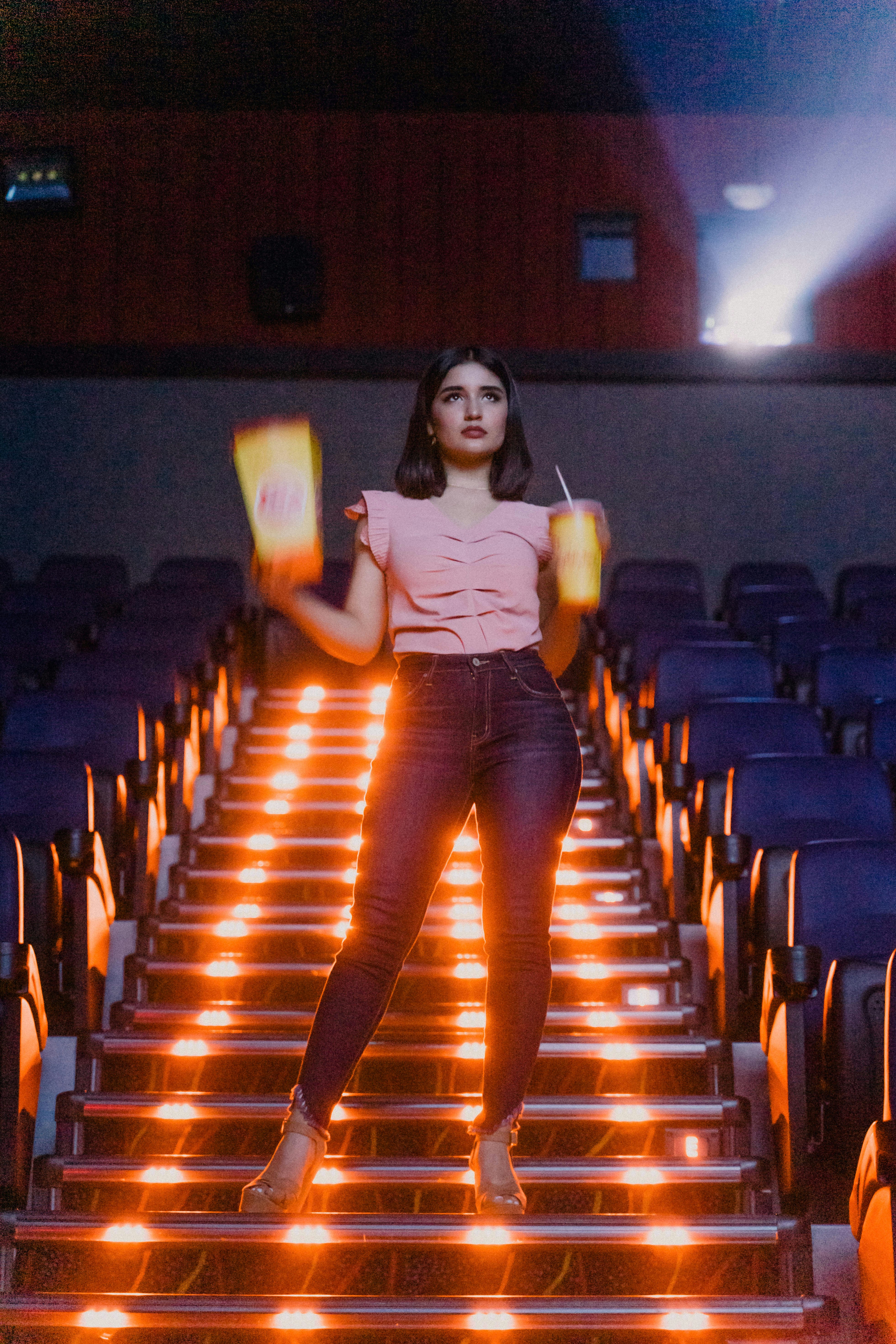 A woman stands on illuminated stairs in a cinema, holding a popcorn box and a drink. Rows of empty seats are visible in a dimly lit auditorium with a spotlight casting light from above.