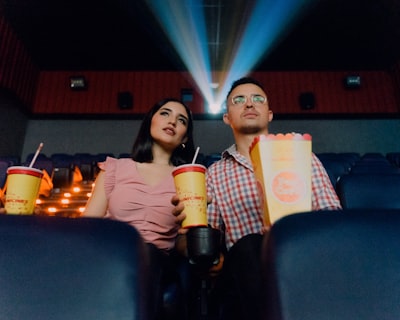 2 women sitting on blue leather chair holding white and red plastic cups popcorn string teams background