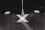 A monochromatic image features an abstract pattern of intersecting lines and shapes projected onto a surface. The focus is on a large X shape created by beams of light. Additional geometric patterns, such as squares and lines, are also visible, creating a symmetrical and minimalist design.