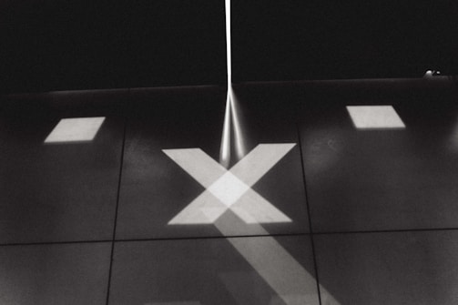 A monochromatic image features an abstract pattern of intersecting lines and shapes projected onto a surface. The focus is on a large X shape created by beams of light. Additional geometric patterns, such as squares and lines, are also visible, creating a symmetrical and minimalist design.