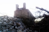 A large industrial building with two tall towers looms in the background, partially obscured by a pile of debris and fallen trees in the foreground. The sky is bright and the overall atmosphere appears desolate and abandoned.