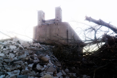 A large industrial building with two tall towers looms in the background, partially obscured by a pile of debris and fallen trees in the foreground. The sky is bright and the overall atmosphere appears desolate and abandoned.