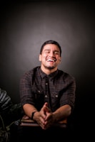 A person is laughing heartily, wearing a dark plaid shirt while sitting indoors. The background is a soft, dark gradient with some green foliage visible at the lower part. The individual’s hands are clasped together, resting on a book or table.