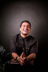 A person is laughing heartily, wearing a dark plaid shirt while sitting indoors. The background is a soft, dark gradient with some green foliage visible at the lower part. The individual’s hands are clasped together, resting on a book or table.