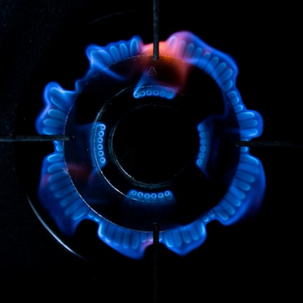 A close-up view of a stove burner with a blue flame. The flame forms a ring around the central burner, displaying an intricate pattern. The black metal grates of the stove are visible above the flame, contrasting sharply with the bright blue color.