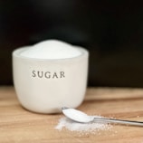 A white ceramic container labeled 'sugar' is filled to the brim with fine white sugar. In front of the container, a stainless steel spoon holds a small amount of sugar, with some spilled sugar scattered on a wooden surface.