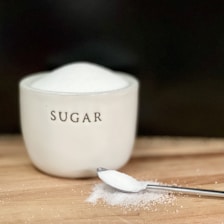 A white ceramic container labeled 'sugar' is filled to the brim with fine white sugar. In front of the container, a stainless steel spoon holds a small amount of sugar, with some spilled sugar scattered on a wooden surface.
