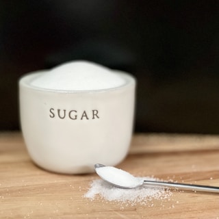 A white ceramic container labeled 'sugar' is filled to the brim with fine white sugar. In front of the container, a stainless steel spoon holds a small amount of sugar, with some spilled sugar scattered on a wooden surface.