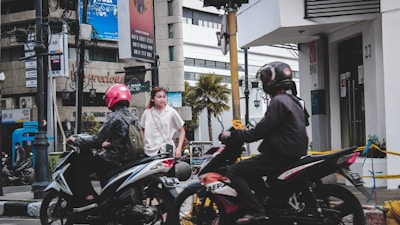 A city street with two motorcyclists wearing helmets passing by a pedestrian. The background includes a modern building with a palm tree and advertisements, along with visible store signs and a bank entrance marked with 'BNI'. The scene captures a lively urban setting with traffic and pedestrians.