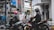 A city street with two motorcyclists wearing helmets passing by a pedestrian. The background includes a modern building with a palm tree and advertisements, along with visible store signs and a bank entrance marked with 'BNI'. The scene captures a lively urban setting with traffic and pedestrians.