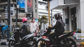 A city street with two motorcyclists wearing helmets passing by a pedestrian. The background includes a modern building with a palm tree and advertisements, along with visible store signs and a bank entrance marked with 'BNI'. The scene captures a lively urban setting with traffic and pedestrians.
