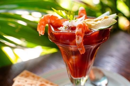 A cocktail glass filled with shrimp cocktail, garnished with shrimp, sliced avocado, and a sprig of fresh herbs. The background features vibrant green leaves, contributing to a tropical ambiance.