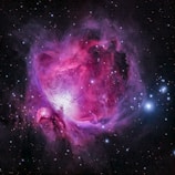 A vibrant and colorful nebula with swirling clouds of pink, purple, and blue hues set against a backdrop of deep space. Numerous stars are scattered throughout, with a bright concentration of light at the center, hinting at a star formation region.