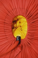 A close-up view of a drawstring bag, predominantly red fabric with a circular opening revealing yellow material inside. The drawstring is thick and red, with a black plastic cinch to secure it.