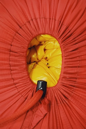 A close-up view of a drawstring bag, predominantly red fabric with a circular opening revealing yellow material inside. The drawstring is thick and red, with a black plastic cinch to secure it.