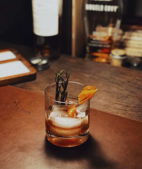 A glass filled with a cocktail, garnished with a sprig of rosemary and a slice of orange peel, placed on a leather surface. The setting includes blurred bottles and a warm, wooden background, suggesting a bar or lounge environment.
