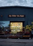 A storefront with the sign 'The Indoor Sun Shoppe' is visible, featuring a large window displaying potted plants and greenery. The building's exterior is a muted gray-blue color, surrounded by various plants and foliage, creating a cozy, inviting atmosphere.