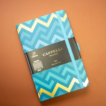 A notebook with a vibrant geometric design featuring zigzag patterns in shades of blue and gold. The cover has a black label with gold lettering that reads 'Castelli Milano Oro Collection'. A matching blue elastic band encircles the notebook, set against a muted brown background.