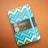 A notebook with a vibrant geometric design featuring zigzag patterns in shades of blue and gold. The cover has a black label with gold lettering that reads 'Castelli Milano Oro Collection'. A matching blue elastic band encircles the notebook, set against a muted brown background.