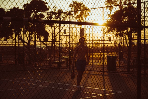 Sunlit outdoor paddle tennis court with a player confidently walking, feathers fluttering gently in the breeze.
