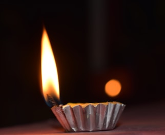 A small flame set in a metallic holder, possibly a diya, cast against a dark background. The flame is bright with shades of yellow and orange, creating a warm glow. A blurred circular light can be seen in the background, enhancing depth.