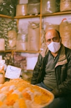 A man wearing a face mask is sitting in a shop with shelves filled with bags of various goods in the background. In the foreground, there is a display of vibrant colored spices or powders.
