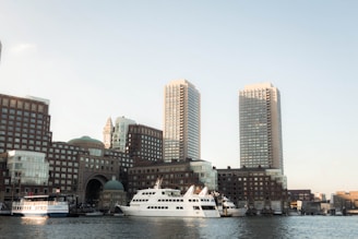 A waterfront cityscape featuring two prominent high-rise buildings surrounded by other modern architecture. Several boats and yachts are docked along the waterside, with clear skies in the background.